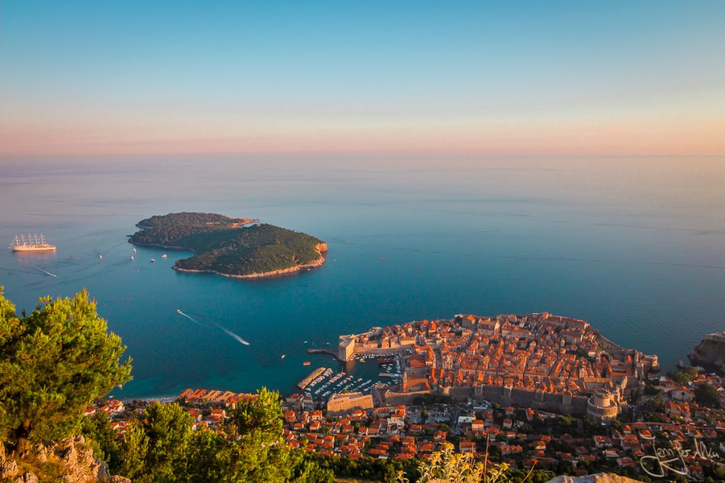 Dieses Bild zeigt die Aussicht vom Brdo Srd. Man blickt über die Stadt Dubrovnik und die Insel Lokrum. Das Meer ist schier endlos und die Sonne geht gerade unter.