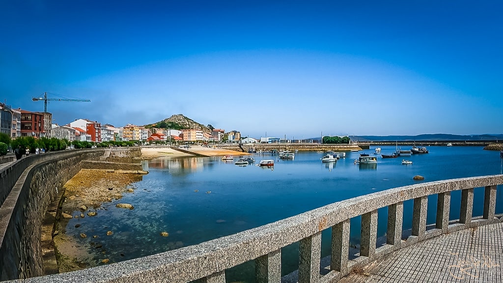 Blick auf den Hafen von Muxía an der Costa da Morte mit ruhigem Wasser, kleinen Booten und der Uferpromenade bei klarem Himmel.