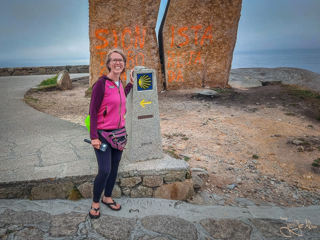 Pilgerin Jennifer Alka steht am 0,0-km-Stein von Muxía, im Hintergrund das Denkmal A Ferida an der Costa da Morte.