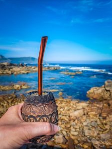 Hand hält einen Mate-Becher vor felsiger Atlantikküste bei Muxia an der Costa da Morte mit Blick auf das Meer.