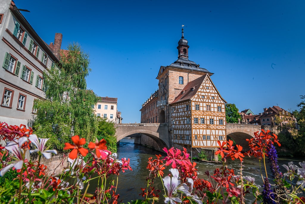 Dieses Bild zeigt das Fachwerk vom Alten Rathaus in Bamberg. Fotografiert vom Geyerswörthsteg mit Blumen im Vordergrund